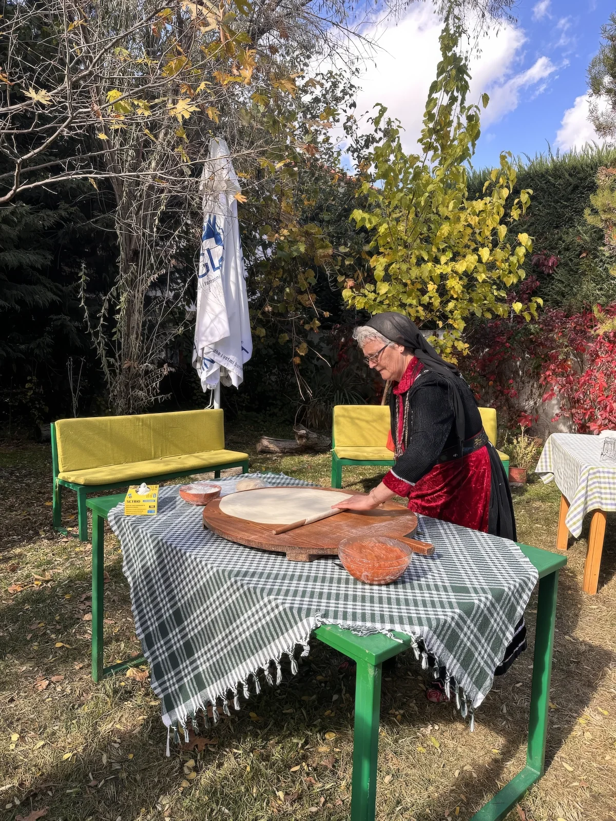 A woman in traditional dress rolling out byrek pastry on a table in a garden