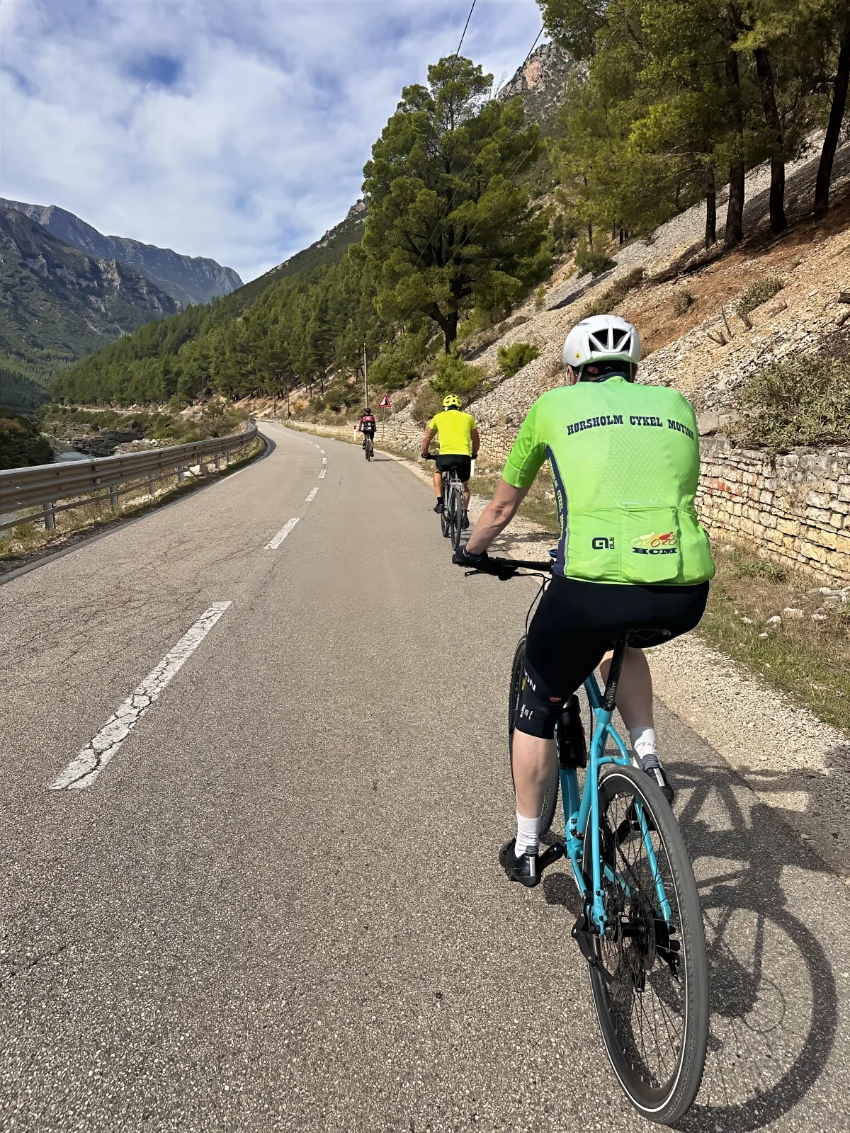 Cyclists in club jerseys riding a quiet road through a wooded valley