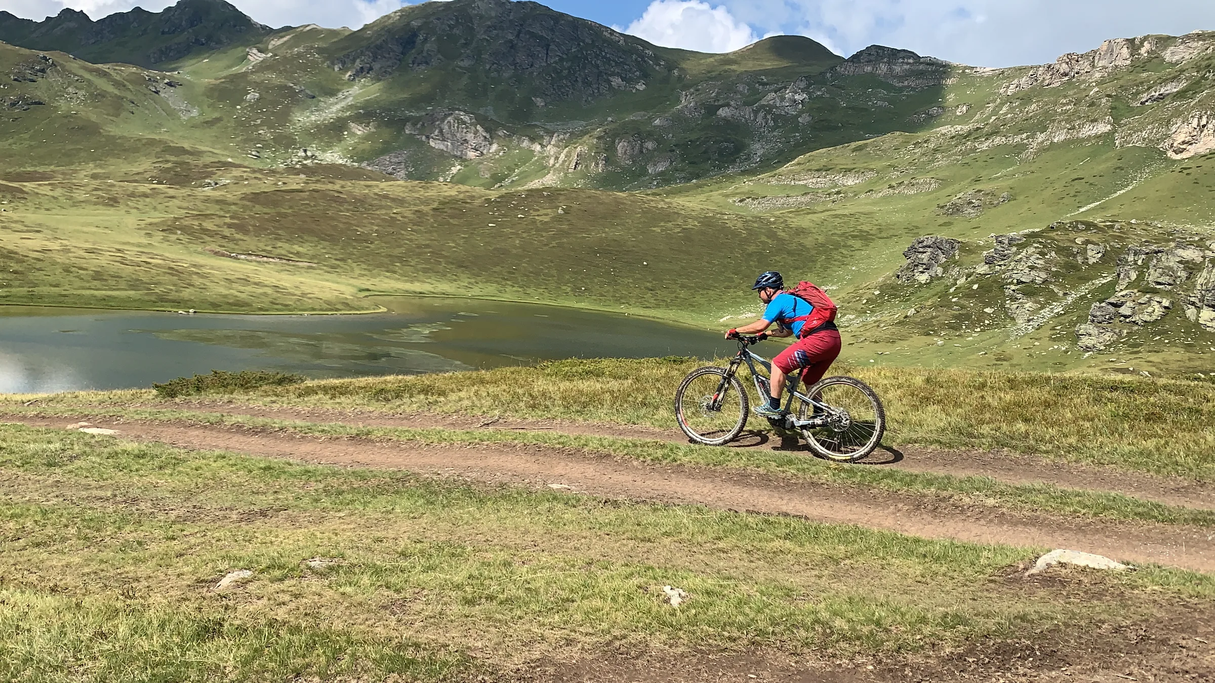 A cyclist riding past a small alpine lake below rocky peaks