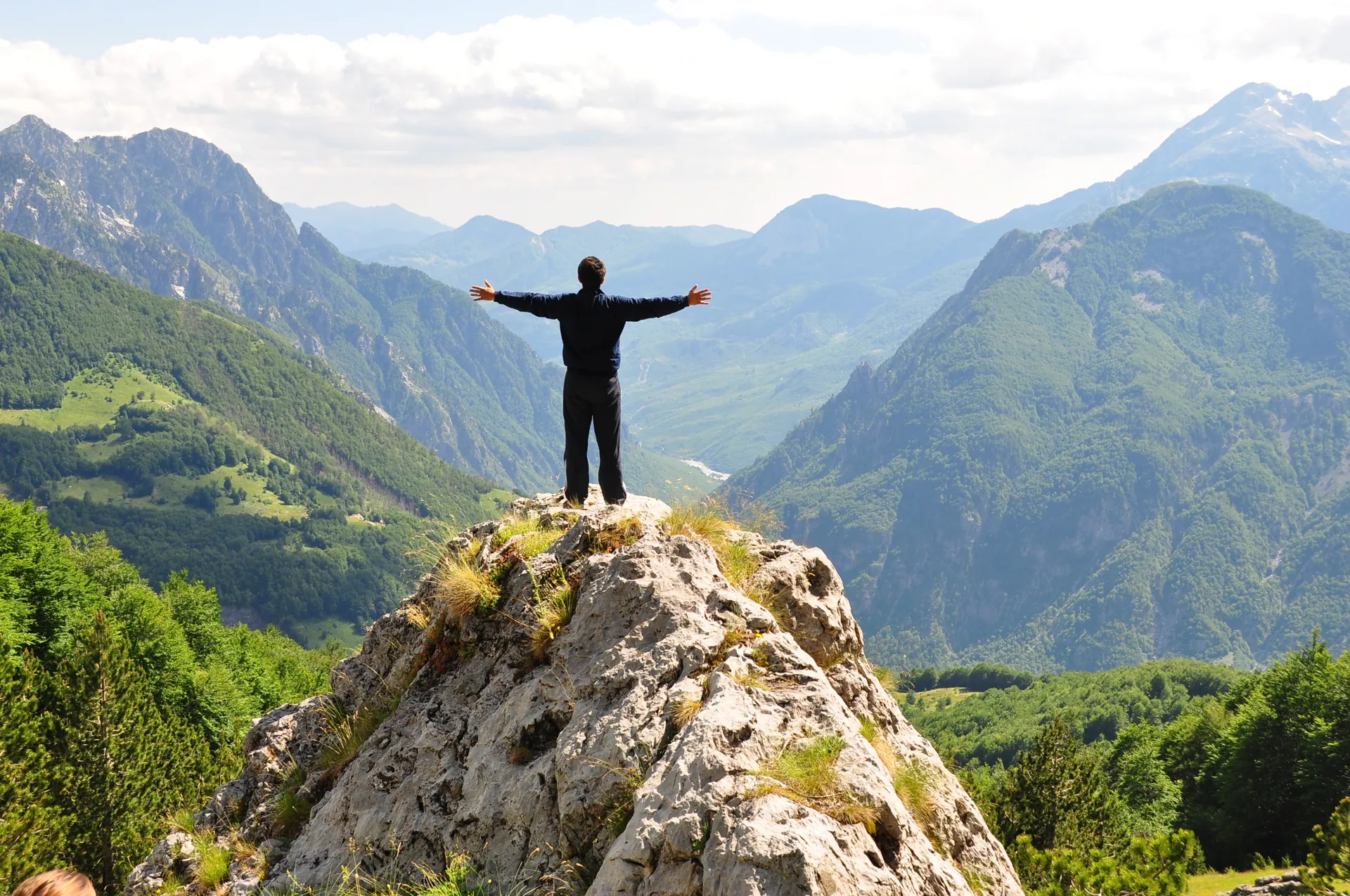 A walker standing with arms wide on a rock above an Albanian valley