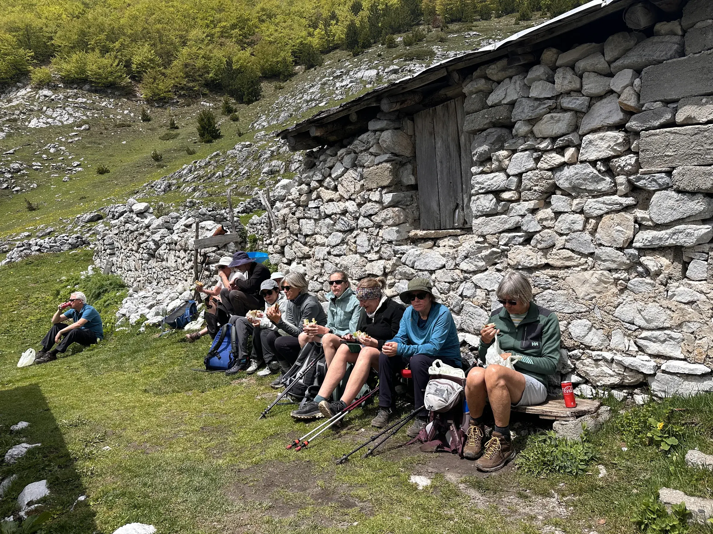 Walkers resting and eating lunch along the wall of a stone shepherd's hut